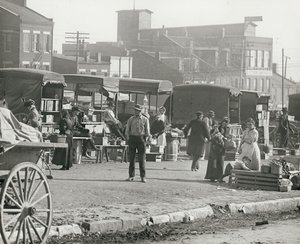 Horizontal, black and white photograph of market stalls and people shopping